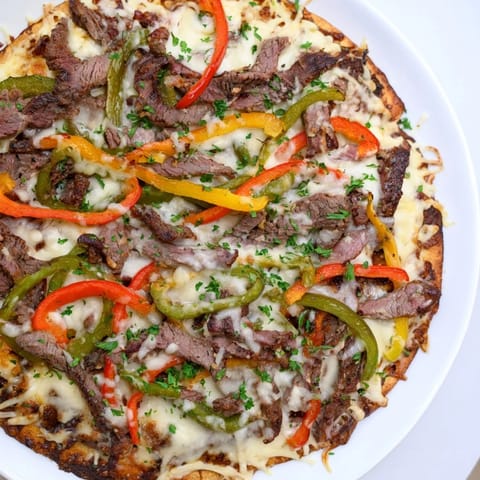 Golden-crusted Philly cheesesteak pizza with visible steak, peppers, and onions ready to be sliced and served.