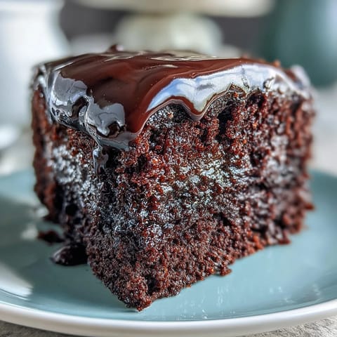 A rich slice of Easy Chocolate Fudge Cake served on a dessert plate, topped with fresh raspberries and a dollop of whipped cream.